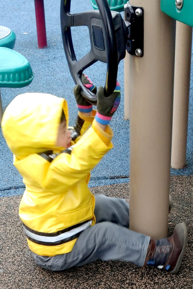 Elliot sitting on the ground, trying to turn the steering wheel above his head at the park